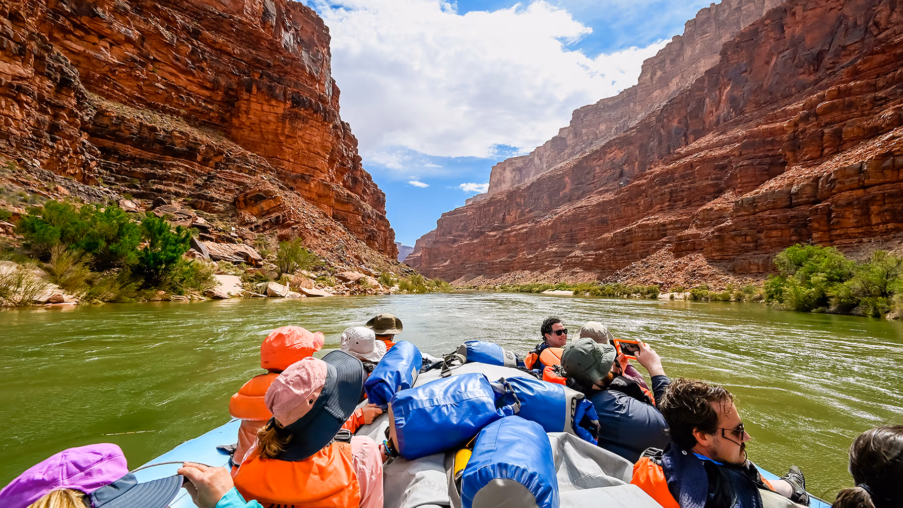 Group of people wearing life jackets rafting on a calm river through a rocky canyon with red cliffs and green bushes.